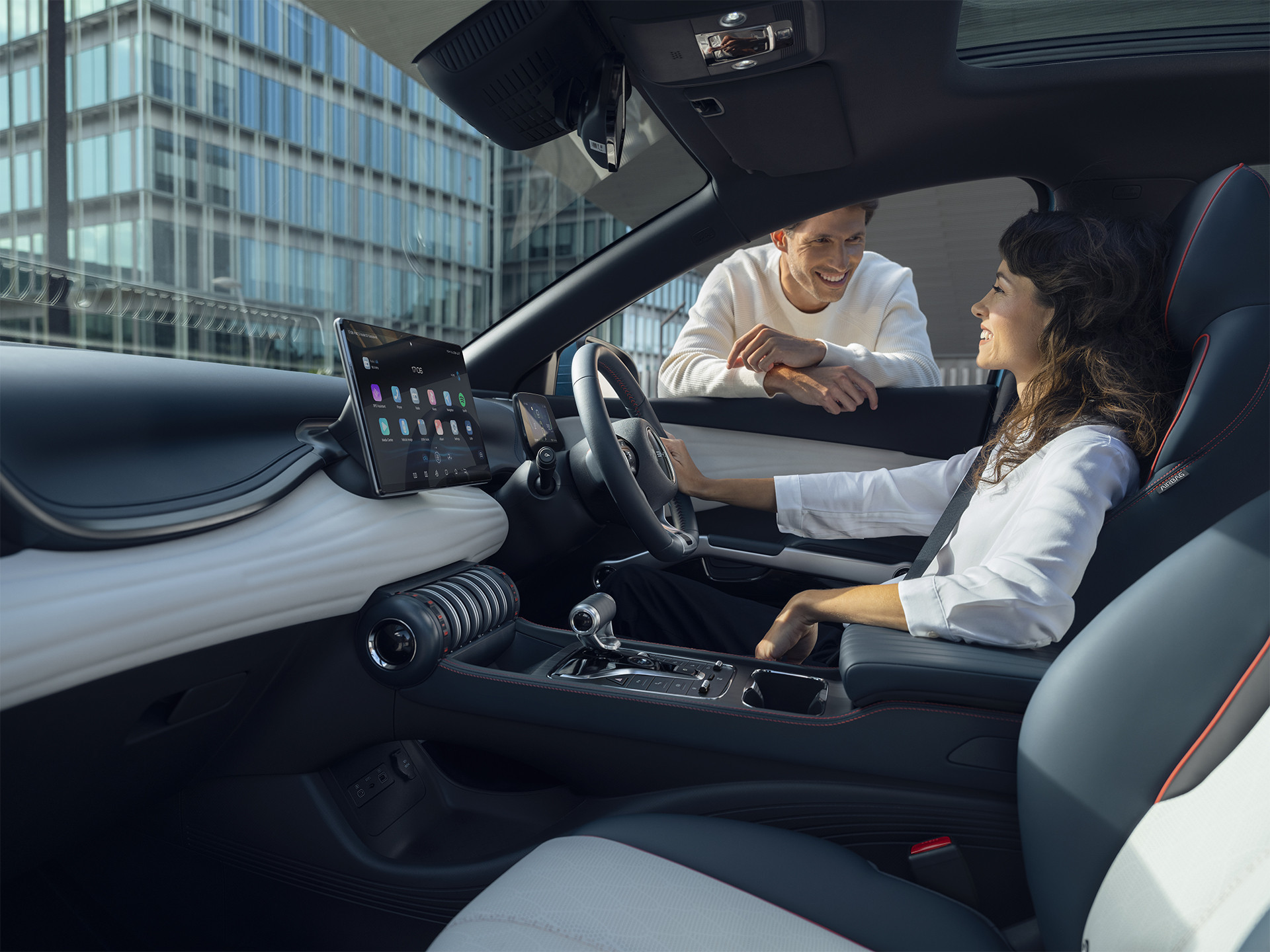 A photograph of a Surfing Blue BYD Atto 3 from the front passenger seat. A woman is sitting in the drivers seat with one hand on the steering wheel. The car is stationary. She is having a conversation with a man who is leaning on the open drivers window.