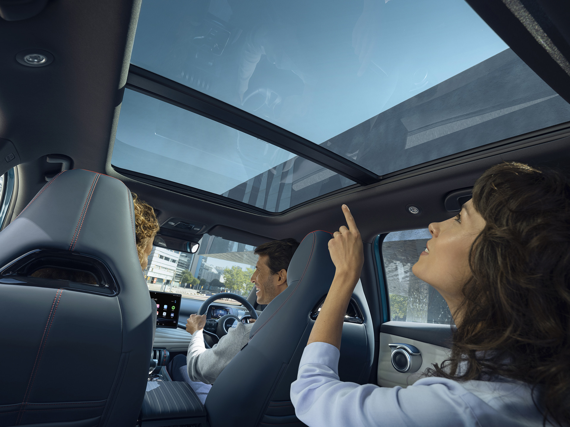 A photograph of a Surfing Blue BYD Atto 3 from the perspective of a rear passenger. Two people in the front are having a conversation. A woman in the rear passenger seat is pointing and looking at the electric sunroof.