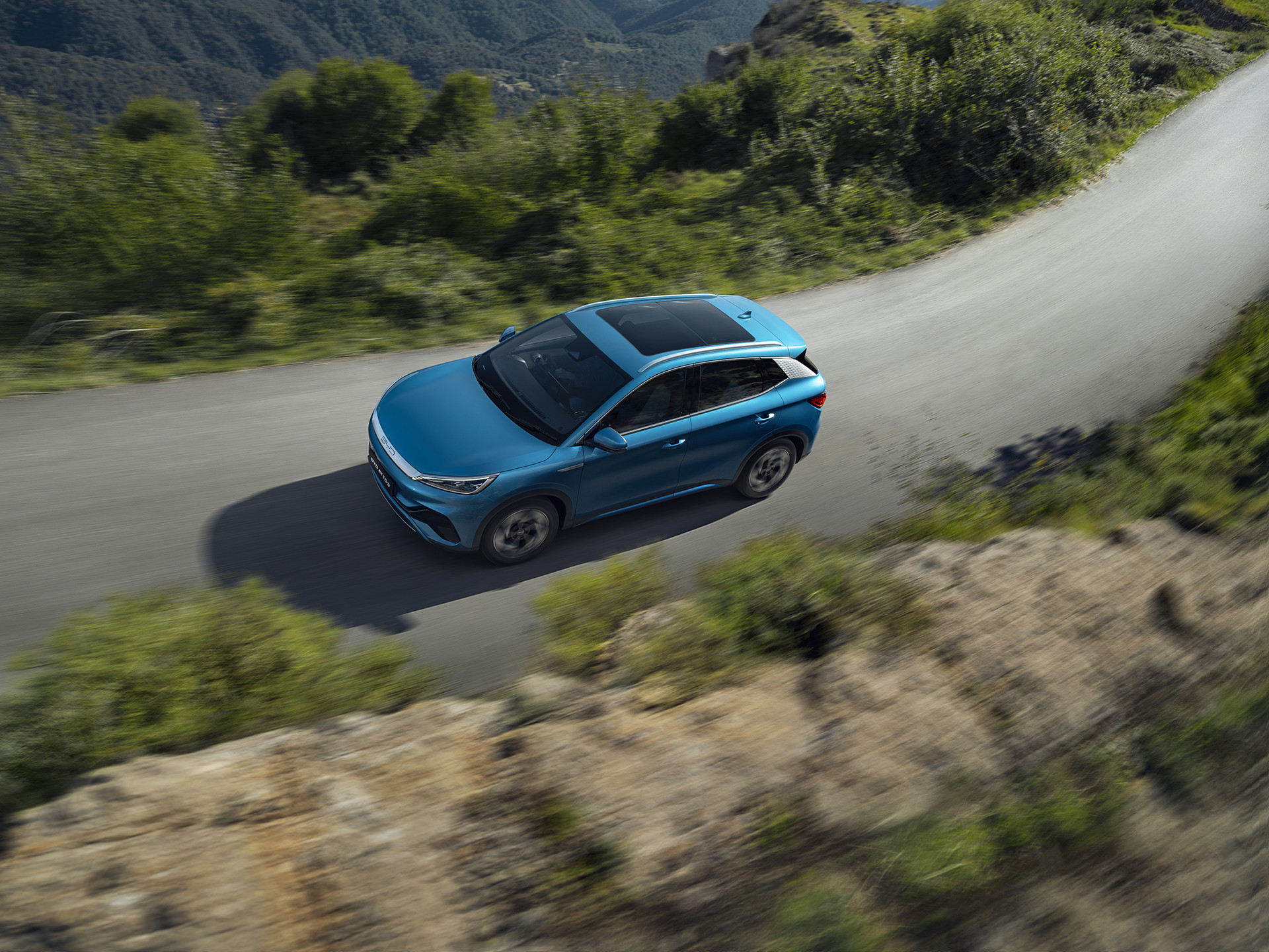 An aerial photograph of a Surfing Blue BYD Atto 3 driving on a narrow country road near a rocky cliff edge